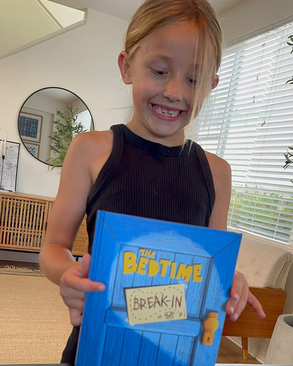 Young girl holding a blue book titled 'The Bedtime Breakin' in a home setting.